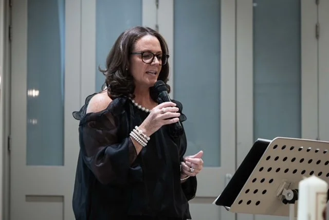 Woman with glasses speaking into a microphone at a music stand with sheet music