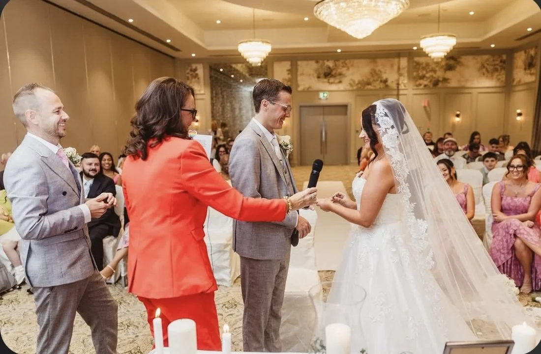 A wedding ceremony with a bride and groom standing in front of an officiant, exchanging rings in a decorated banquet hall with seated guests watching and smiling.
