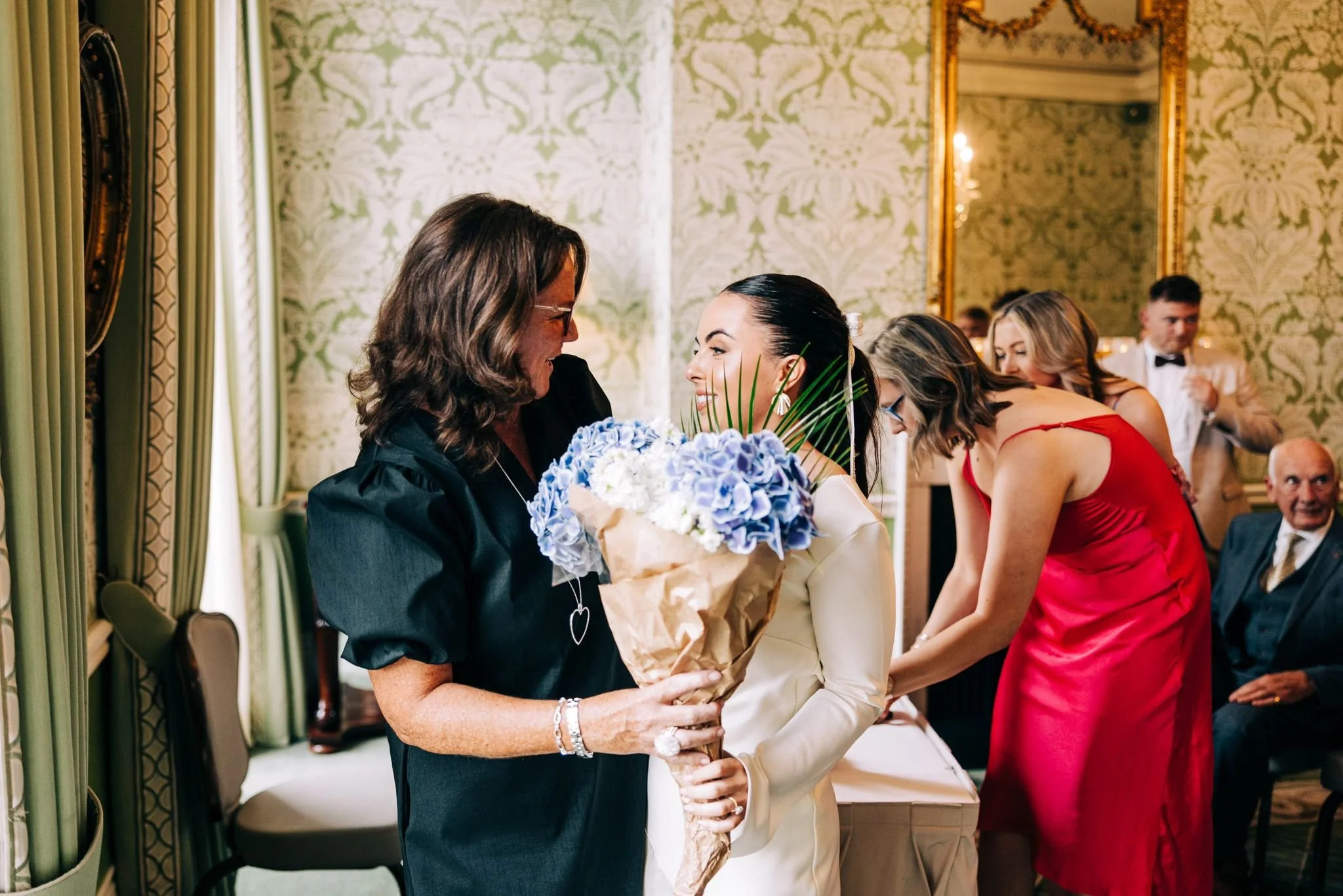 A woman in a white dress holding a blue and white flower bouquet is smiling and talking to an older woman in a black dress, inside a decorated room with ornate wallpaper and mirrors, with other women and men in formal attire in the background.