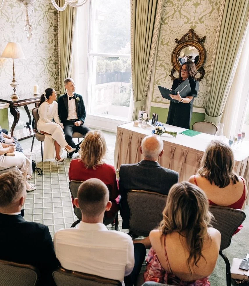 Wedding ceremony with a bride and groom seated while a woman reads from a book, surrounded by guests in a decorated room with large windows and green curtains.