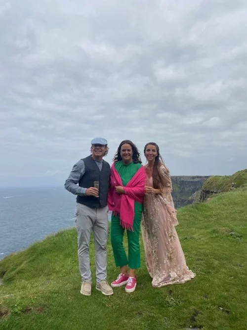 Three women standing together on a grassy cliff overlooking the ocean under a cloudy sky, smiling for a photo.
