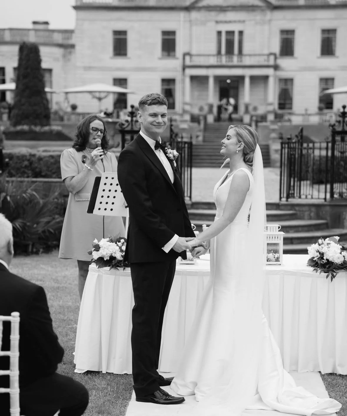 A black and white photo of a wedding ceremony taking place outdoors, with a bride and groom holding hands and exchanging vows, an officiant reading from a paper, all in front of a grand building.