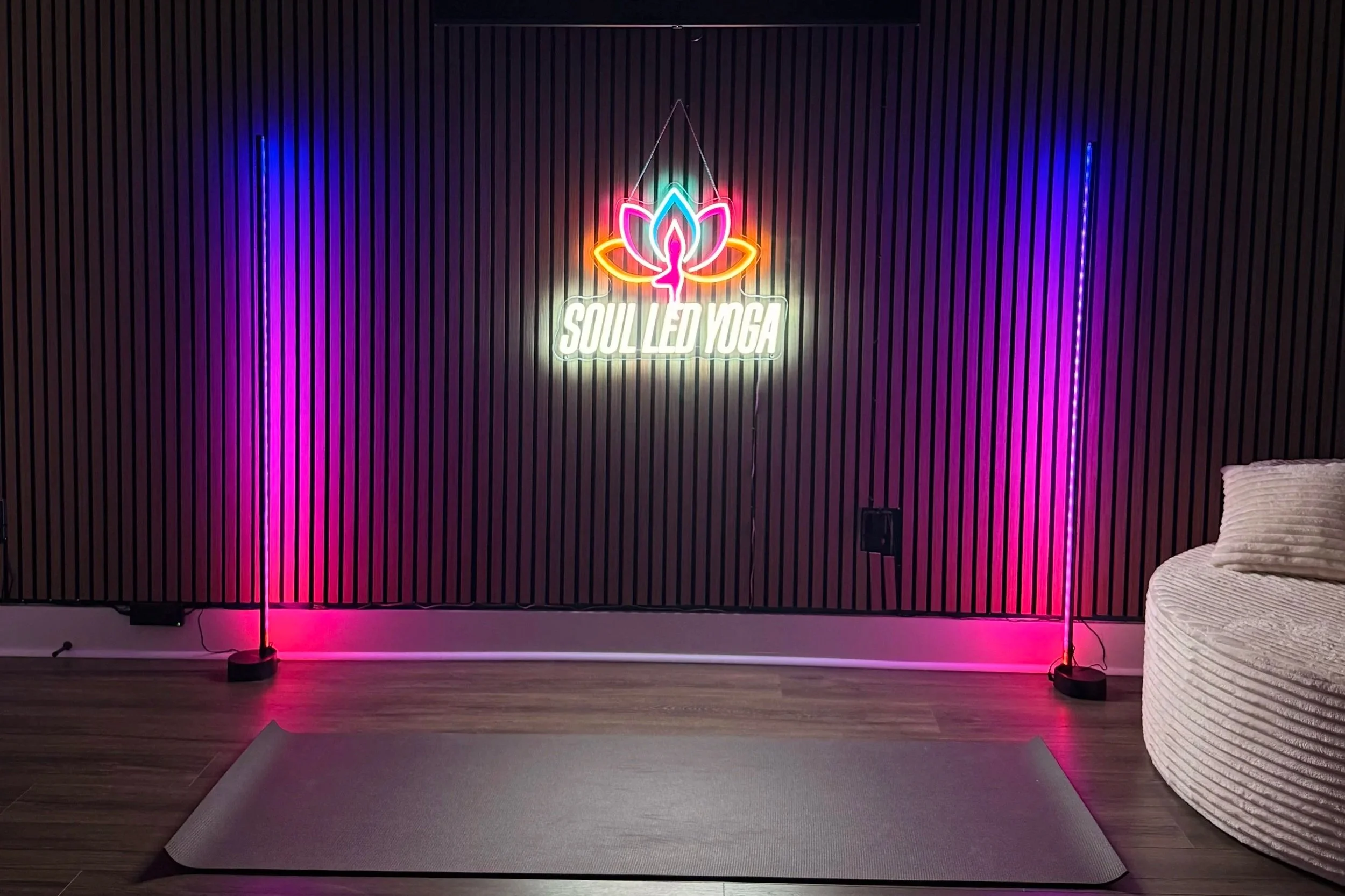 Neon sign on a black wooden wall with a lotus flower and a silhouette of a person in a yoga pose, reading 'Soul Led Yoga'. Vertical LED light strips on each side emit pink and blue light. Gray yoga mat on a wooden floor in front of the wall, with part of a white, textured, circular cushioned seat visible on the right.