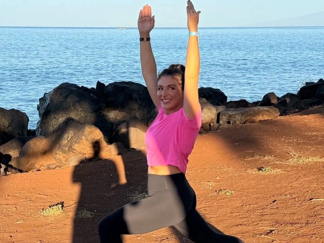 A woman in a pink top and gray leggings performs a yoga pose by the water, with rocky shoreline and ocean in the background; her shadow is cast on the sand.