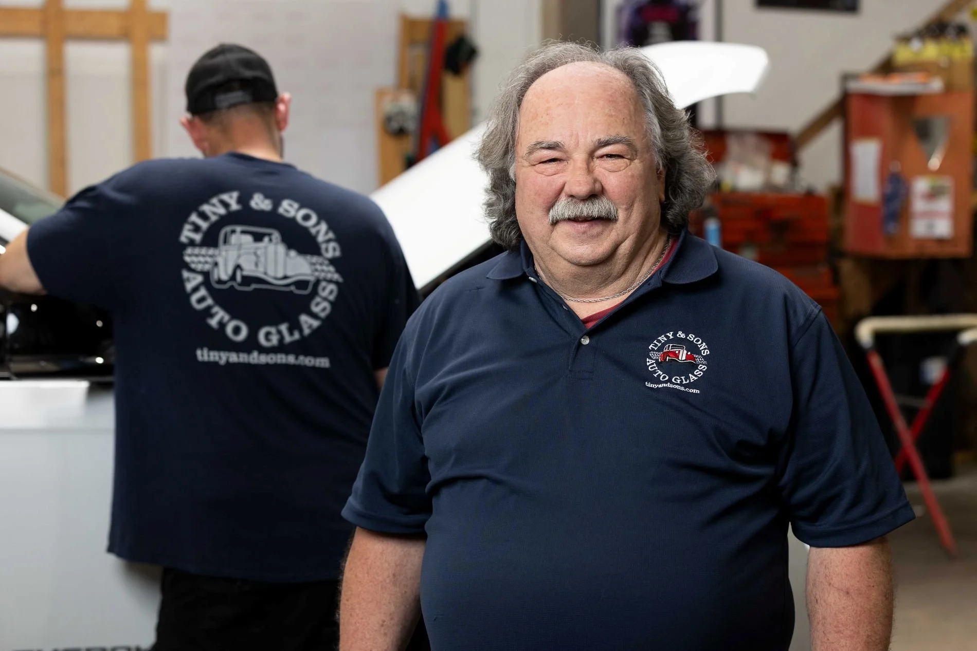 A smiling man with gray hair and a mustache wearing a navy blue polo shirt with the logo 'Tiny & Sons Auto Glass'. In the background, another person with a hat and a similar shirt is working in a garage.