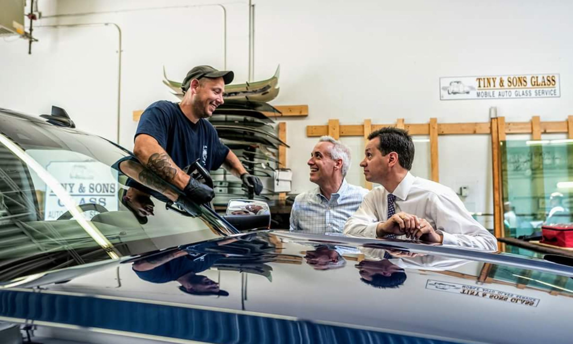 Three men in a garage, two seated and one standing, engaging in conversation. The standing man is smiling and holding a tool, while the seated men listen and look at him. Car windshields and glass panels are visible in the background, along with a sign that reads "Tiny & Sons Glass".