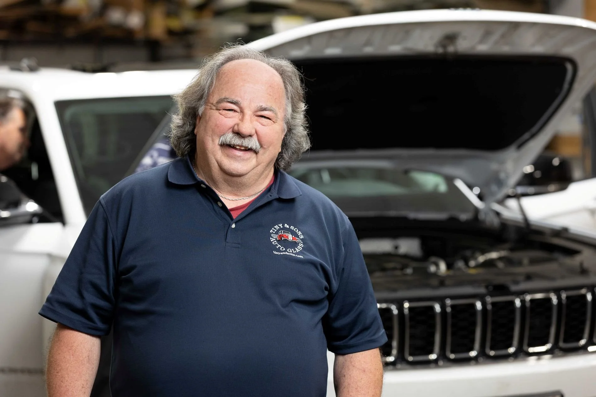 A smiling man with gray hair and a mustache wearing a navy blue polo shirt with a logo, standing in front of a car with its hood open inside a garage or workshop.