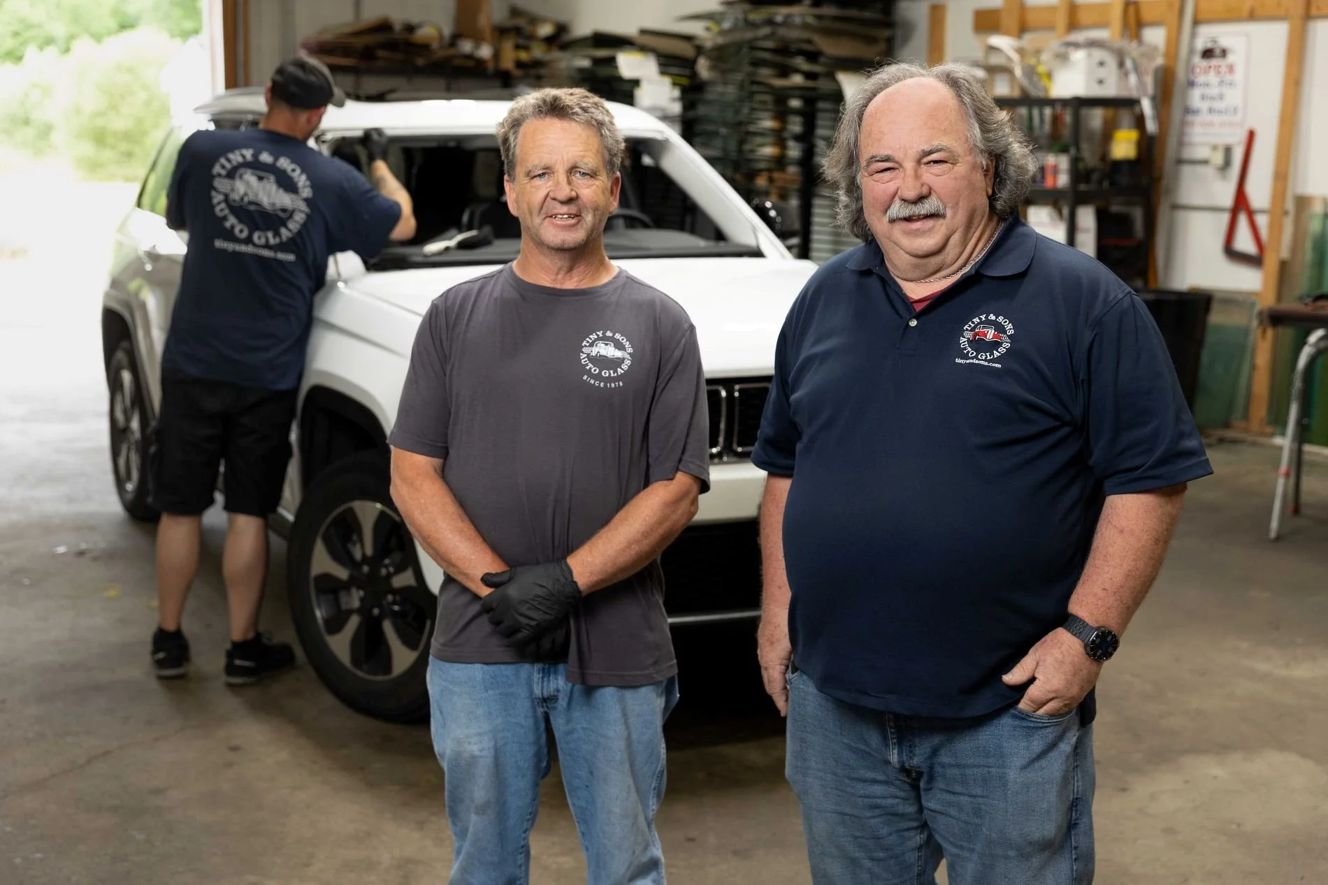 Two men standing in front of a white SUV inside a garage, with a person working on the vehicle in the background. All three are wearing shirts with the logo 'Tiny & Sons Auto Glass.'