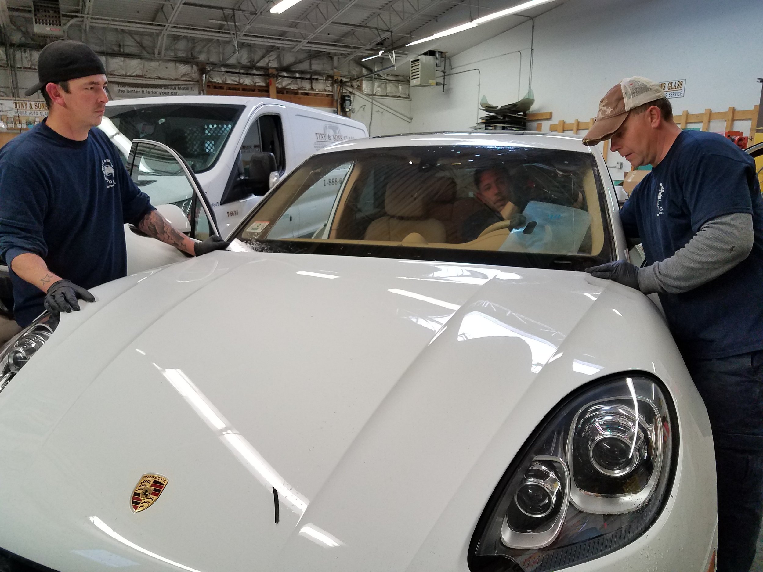 Two men in blue shirts and gloves work on a white Porsche car inside a garage or workshop. One man on the left is touching the car's hood while the other on the right leans on the car's front fender. A man looks through the windshield from inside the car.