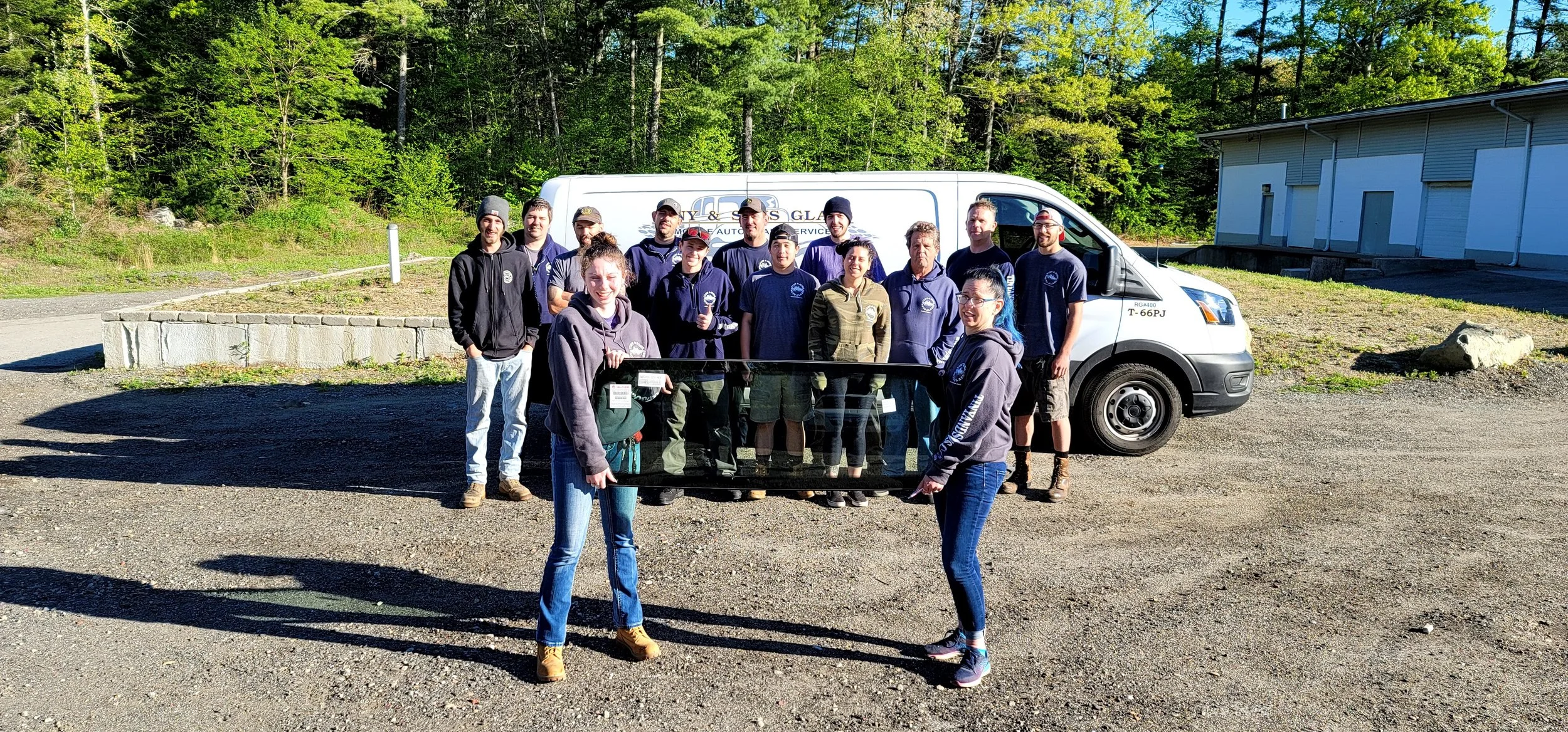 A group of people standing outdoors in front of a white service van and a building, holding a large black framed glass panel, with trees and bright blue sky in the background.