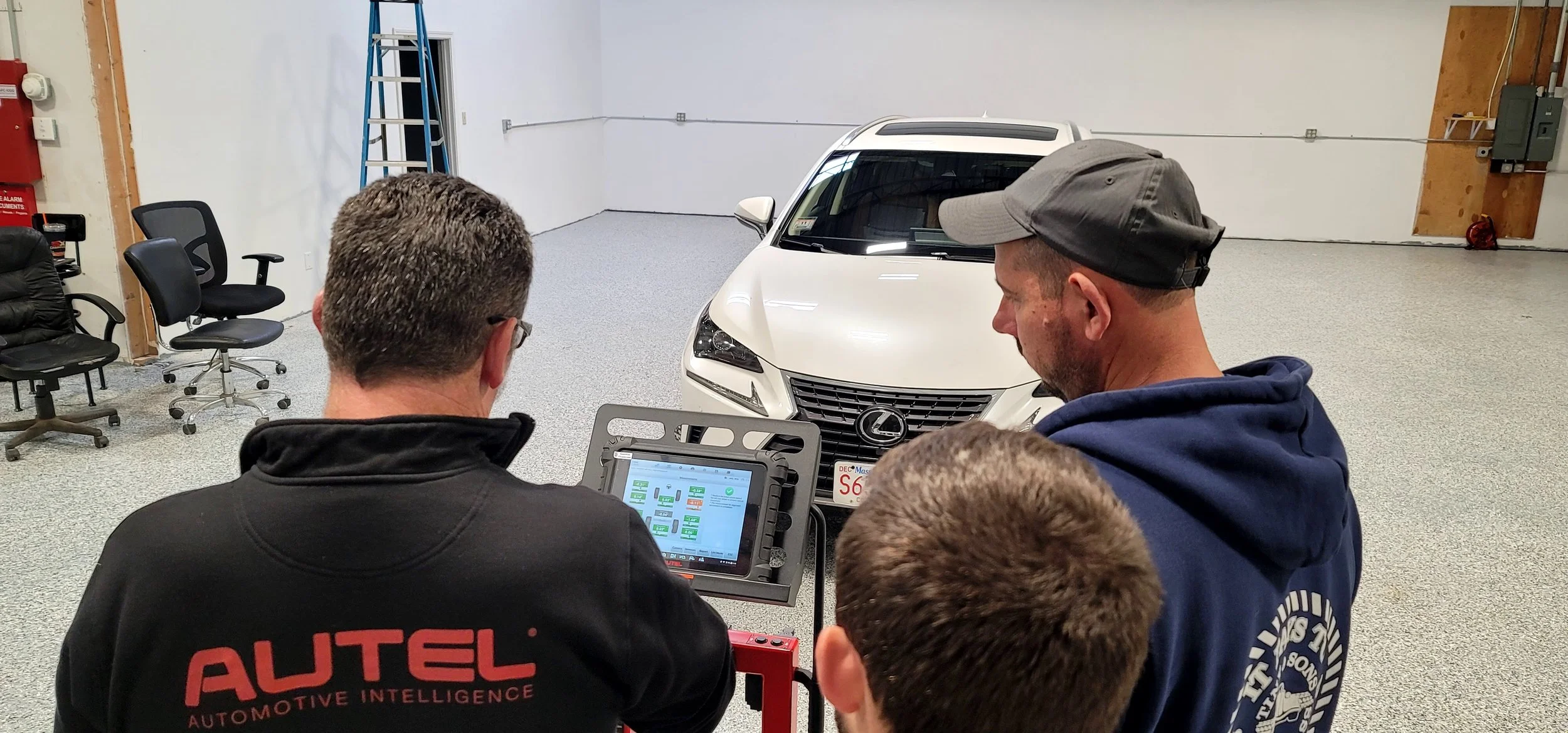 Three men gathered around a diagnostic tablet in a garage with a white Lexus car in the background. Two chairs and a ladder are on the left side. The man with his back to the camera is wearing a black jacket with 'AUTEL AUTOMOTIVE INTELLIGENCE' written on it. The man to his right has a gray cap and a blue hoodie, and a third person is partially visible in front of them.