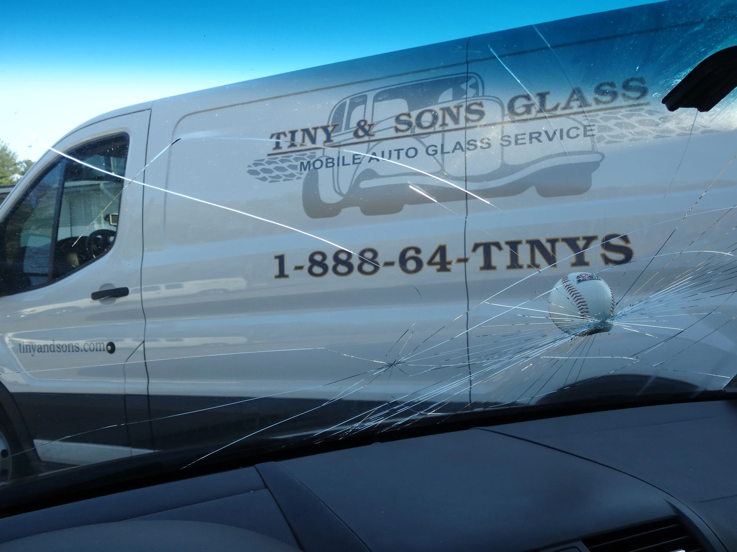View of a white van with a cracked windshield, displaying the logo and contact information for Tiny & Sons Glass, a mobile auto glass service, with a baseball embedded in the crack.