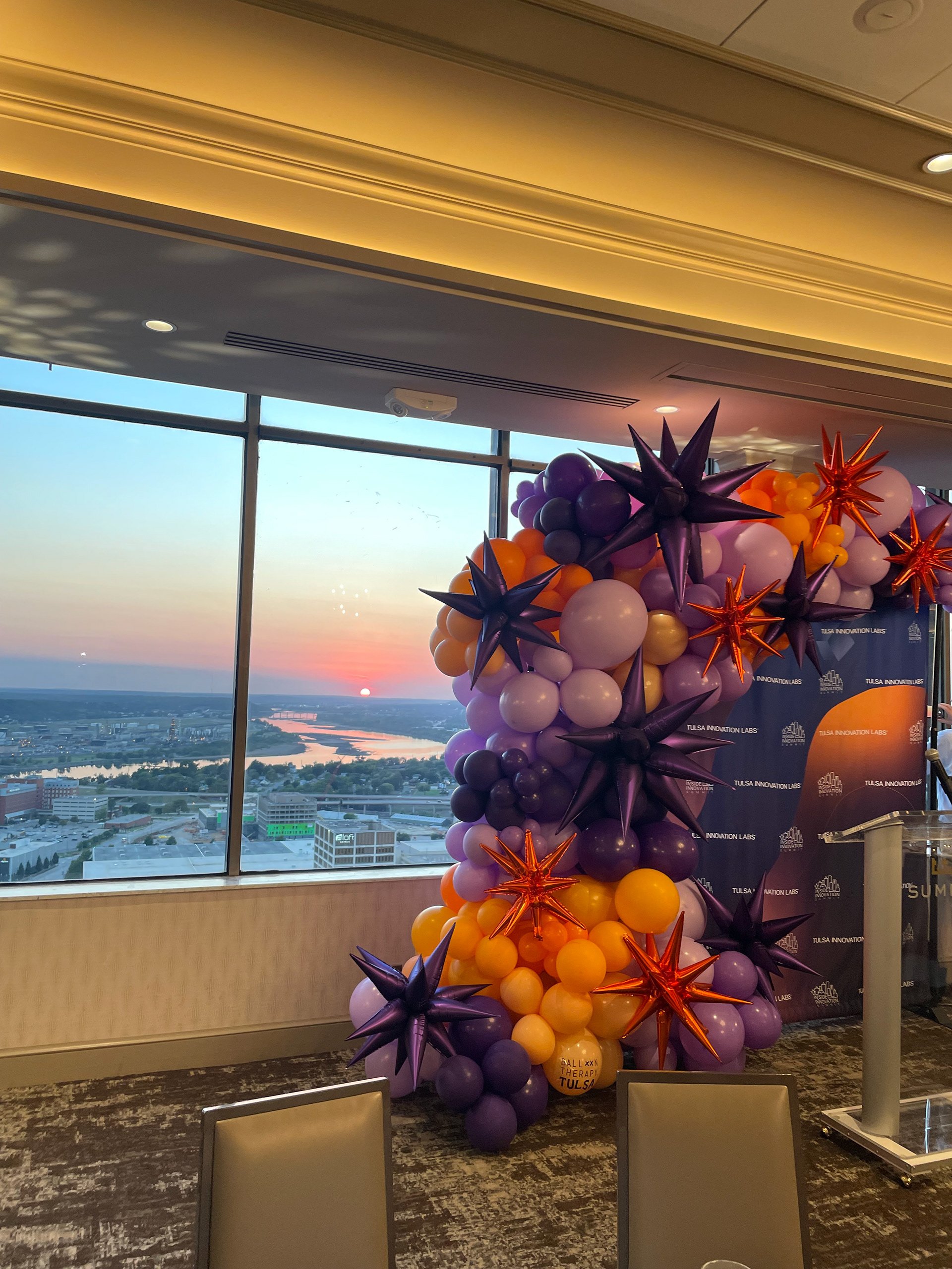 Balloon display consisting of purple, yellow, white, and orange balloons with star-shaped balloons in purple and orange, set indoors with large windows showing a sunset over a cityscape with river.