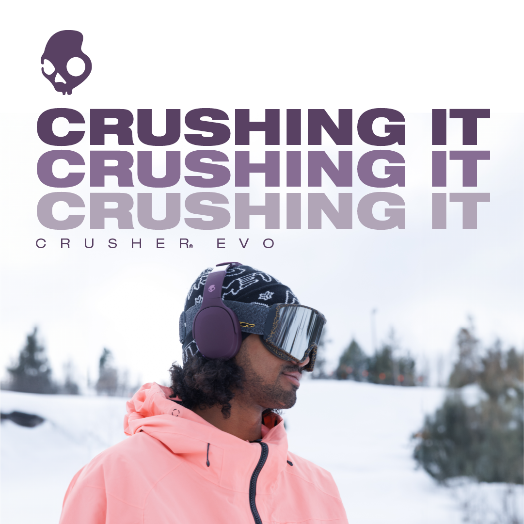 A person in a pink jacket and black patterned headwear wearing purple headphones and ski goggles in a snowy outdoor setting.