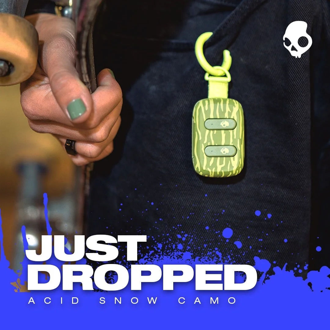 Close-up of a person's hand holding a set of keys with a yellow-green camouflage patterned key fob attached and a dark blue jacket. The image also contains text saying 'Just Dropped' and 'Acid Snow Camo' along with a graphic of a skull in the corner.