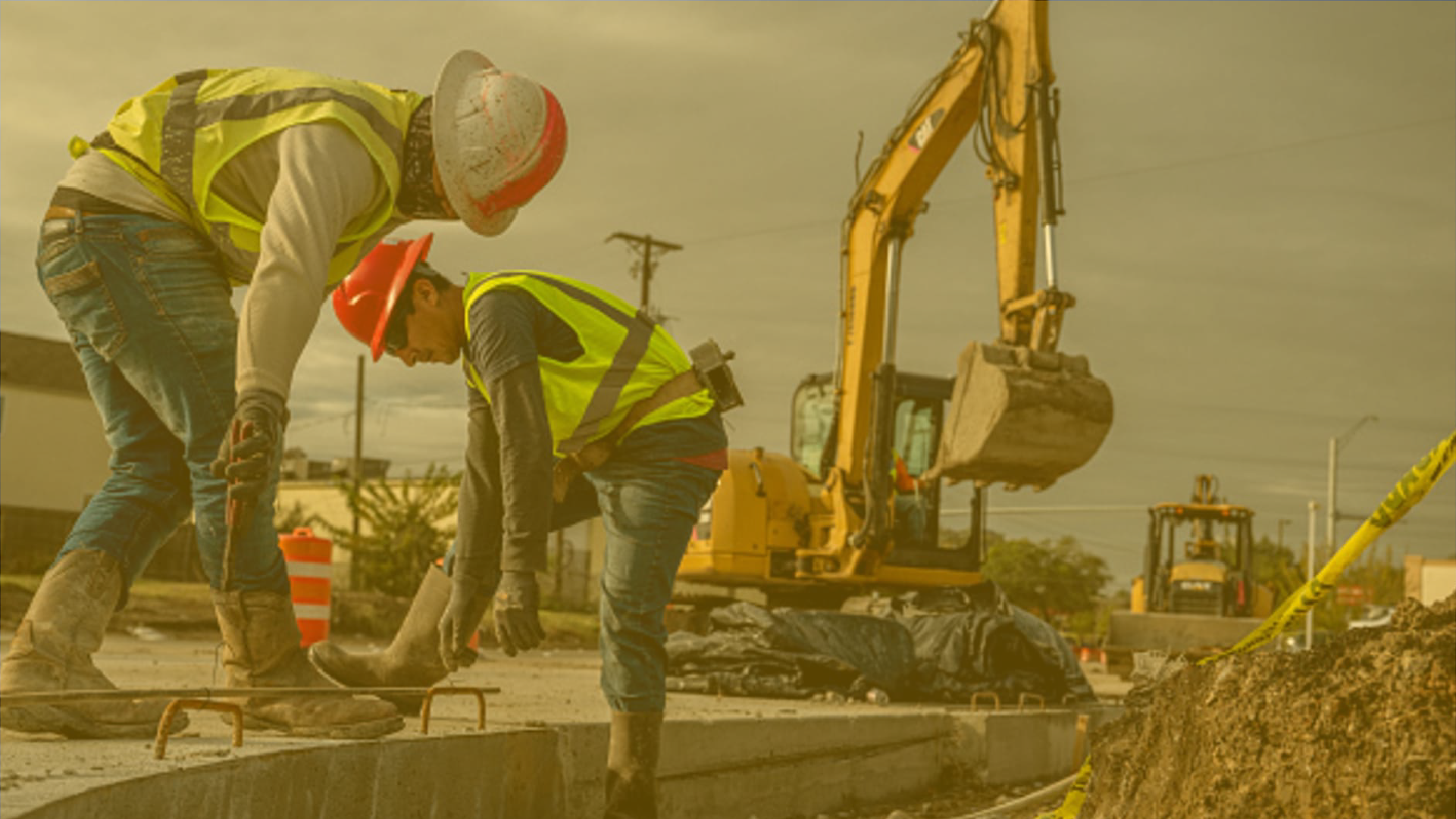 Two construction workers wearing safety vests and helmets working on a road with an excavator in the background and construction cones nearby.