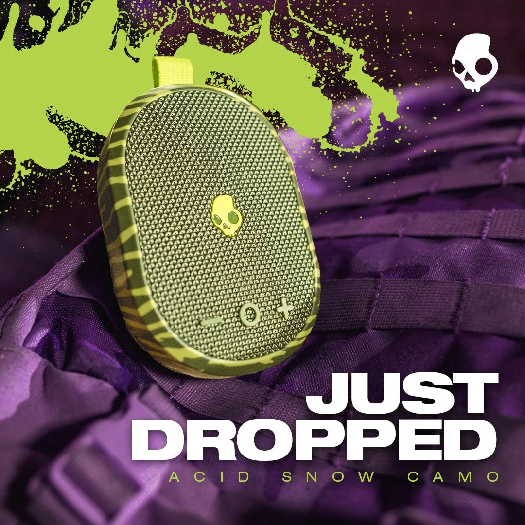 A small electronic device with a skull logo, placed on a purple fabric surface, with a background featuring camouflage patterns and text reading "Just Dropped" and "Acid Snow Camo".