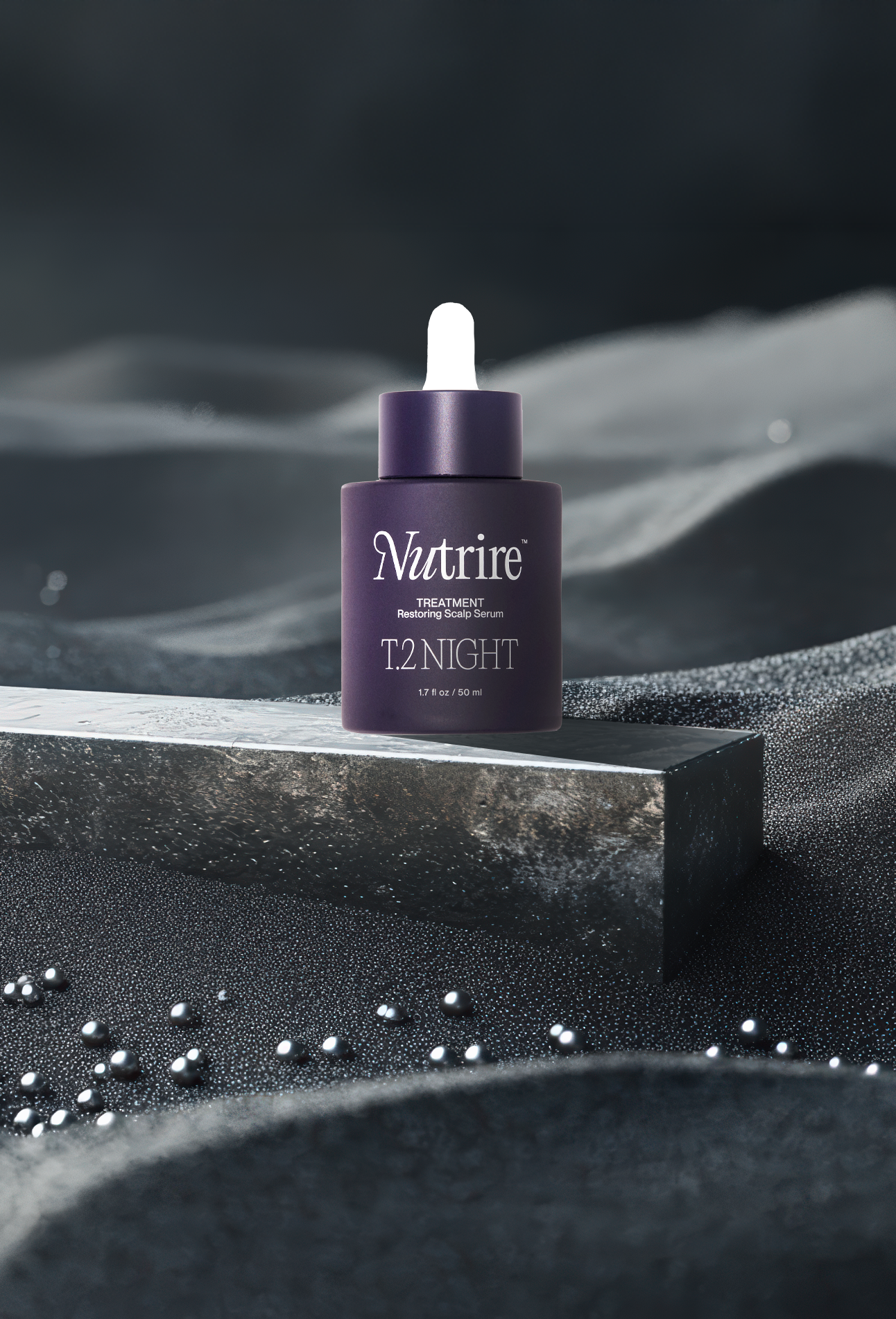 A skincare serum bottle labeled 'Nutrire T.2 Night' positioned on a metallic surface with dark textured materials and scattered small silver beads, illuminated with dramatic lighting.