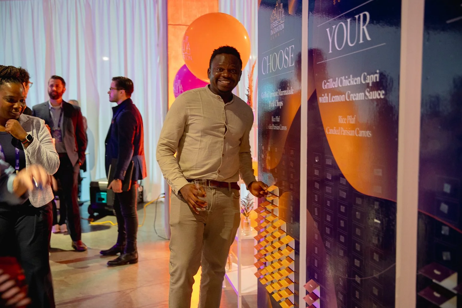 A man in a beige shirt and light pants smiling and holding a glass of drink, standing near a colorful food menu board at a social event with other people in the background.