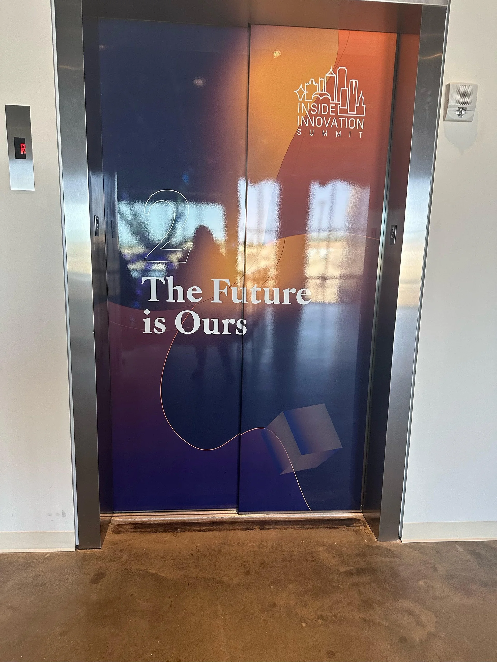 Elevator doors wrapped in promotional material for Inside Innovation Summit, featuring the text "2 The Future is Ours" and a graphic of a city skyline with a book.