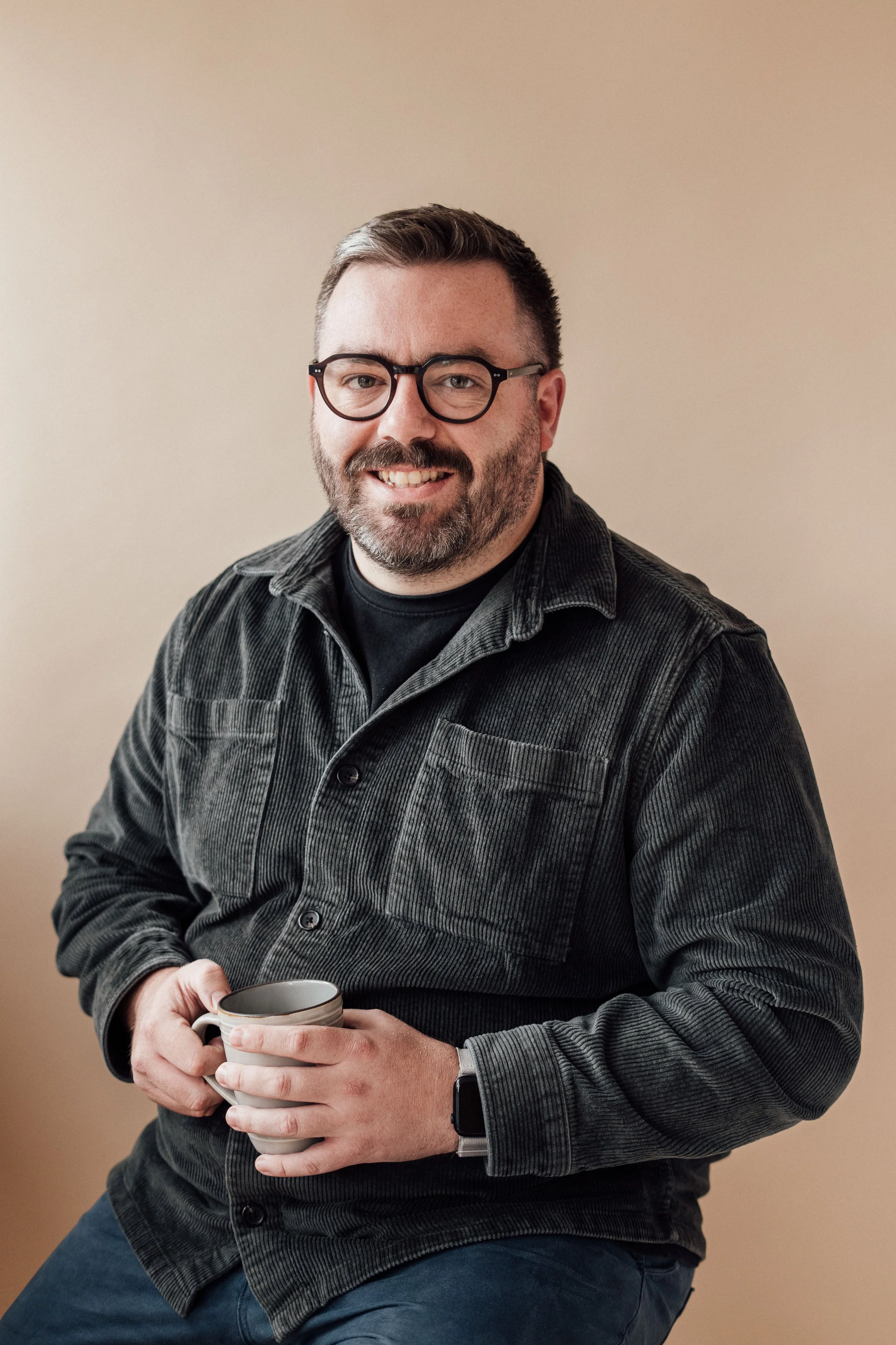 A man with glasses and a beard, smiling, holding a coffee mug, wearing a dark gray corduroy shirt, sitting against a beige background.