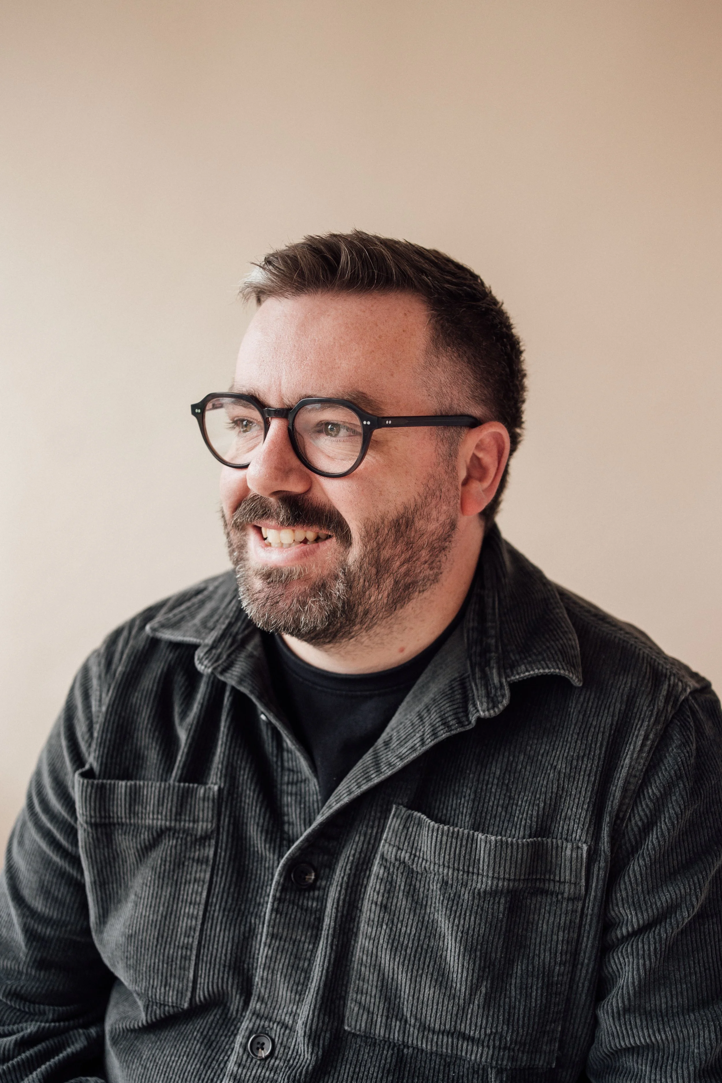 A man with glasses and a beard smiling, wearing a dark button-up shirt, sitting against a plain light-colored wall.