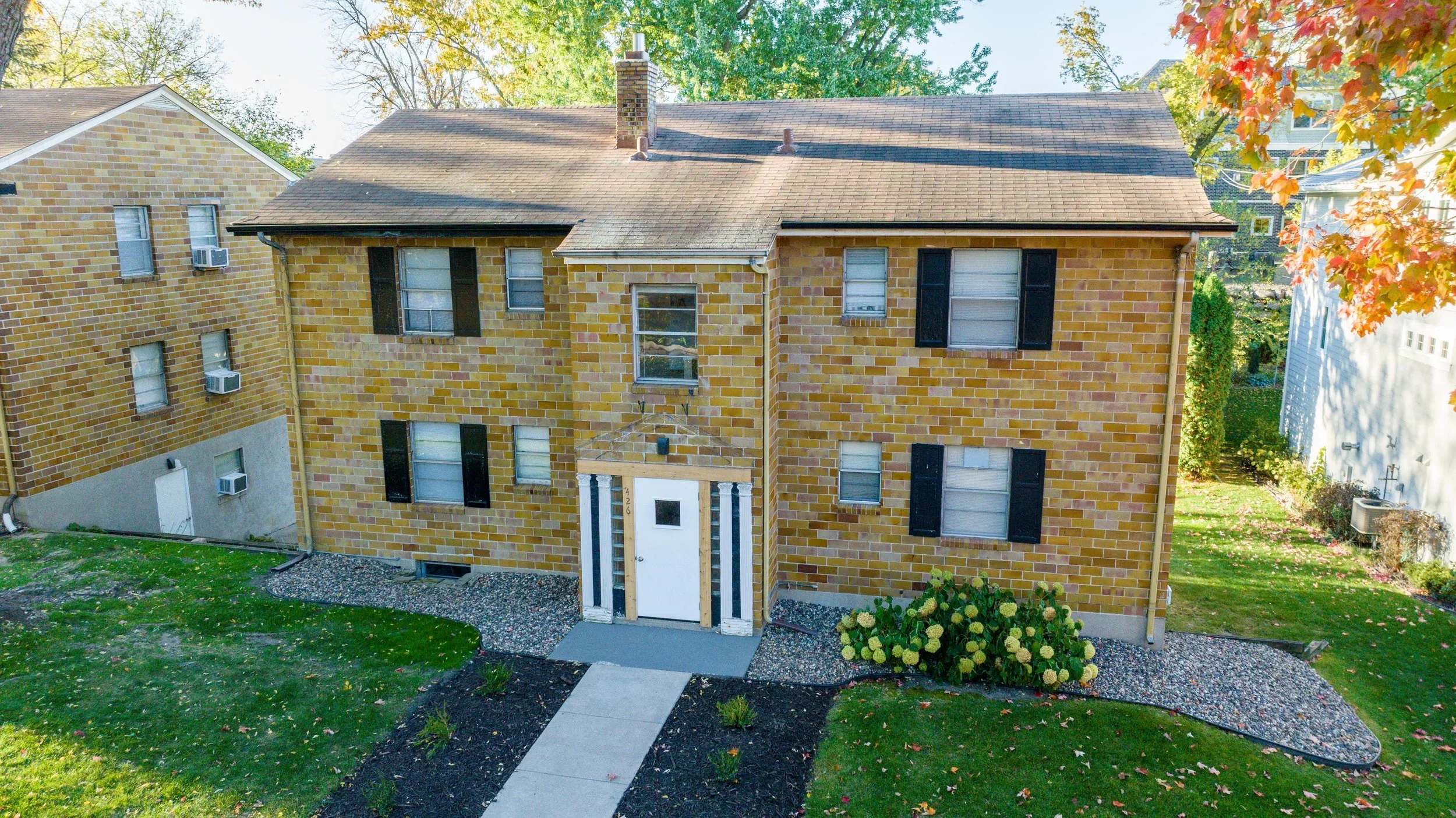An aerial view of a brick apartment building with multiple windows, black shutters, and a white front door with decorative columns. The building is surrounded by a well-maintained yard with green grass, flowering bushes, and landscaped pathways, with trees showing fall foliage.