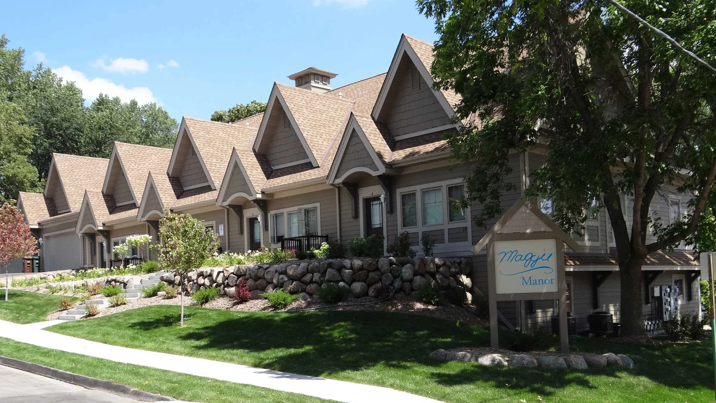 A multi-unit residential building with a sloped roof and dormer windows, surrounded by landscaped greenery and a sign reading "Maggie Manor."