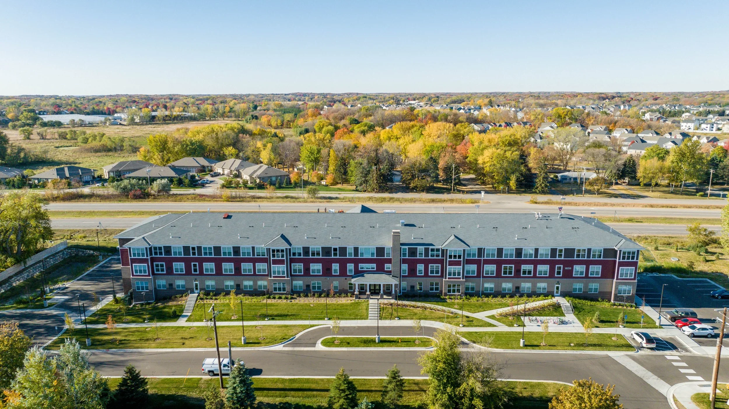 Aerial view of a multi-story apartment building with a parking lot, surrounded by trees and residential neighborhoods under a clear blue sky in autumn.