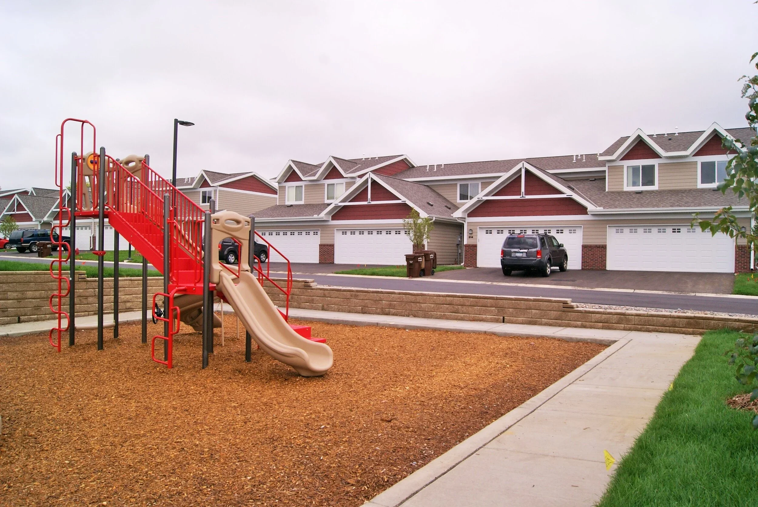 Playground with a slide and climbing structure in a residential neighborhood with townhouses in the background.