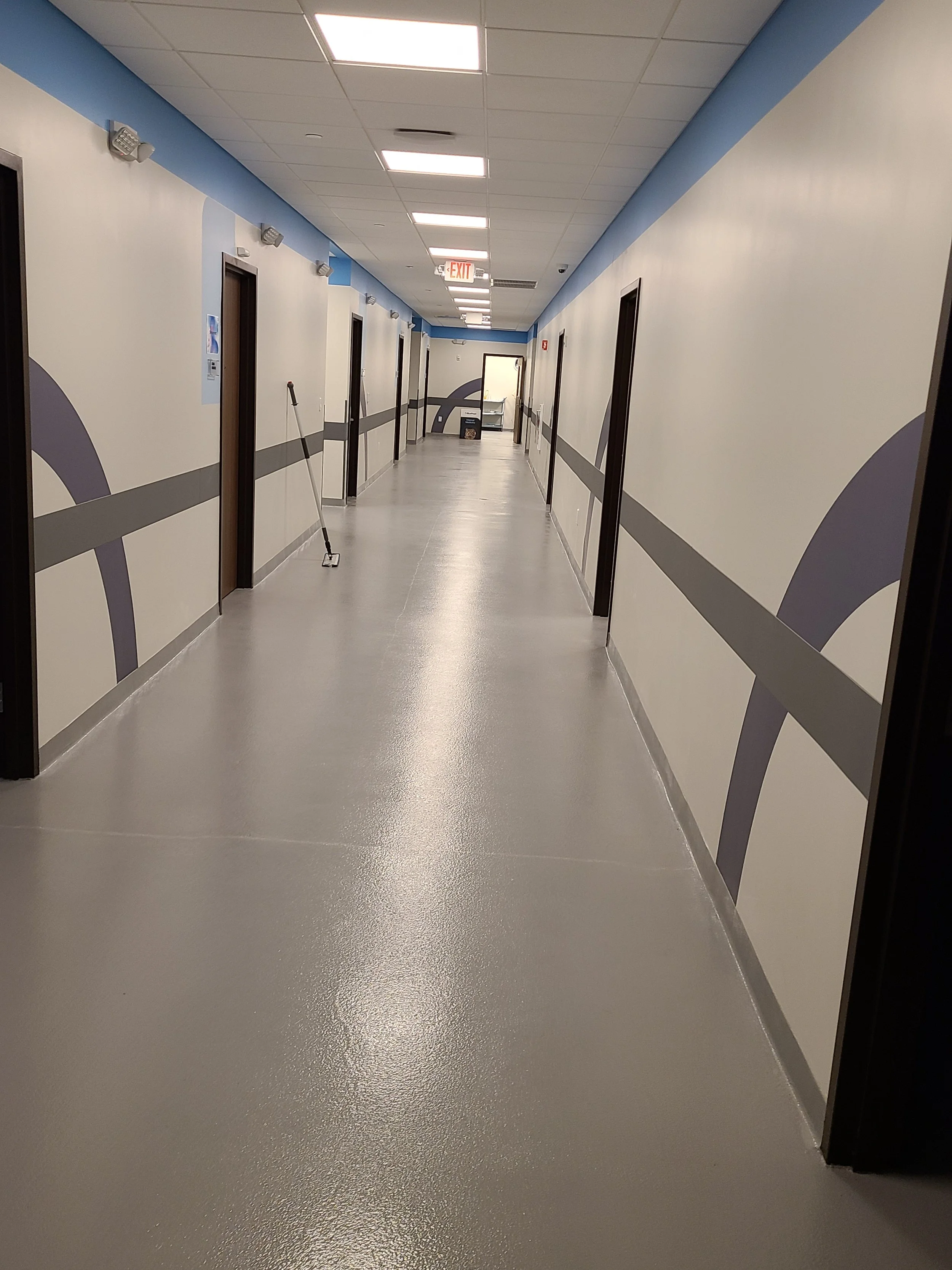 Empty hallway with closed doors on either side, mop leaning against the wall, and exit sign at the end of the corridor.