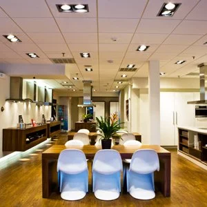 Modern dining area with six white chairs around a wooden table, potted plant, and a contemporary kitchen in the background.
