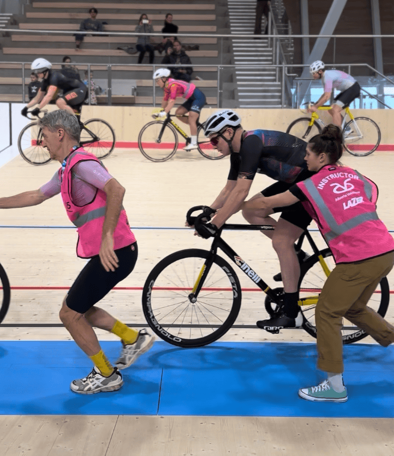 A person on a bicycle being assisted by a staff member during a track cycling event in an indoor velodrome, with other cyclists riding in the background and spectators seated above.