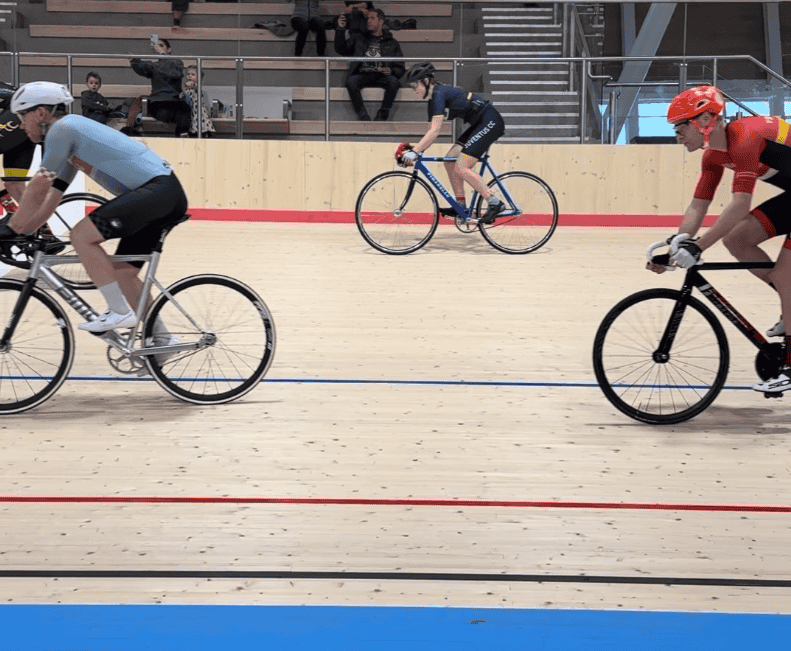 Cyclists racing on an indoor velodrome track with spectators watching from the stands.