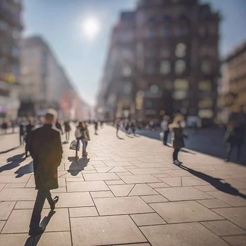 City street scene with pedestrians walking on a sunny day, blurred buildings in the background, and long shadows cast on the pavement.