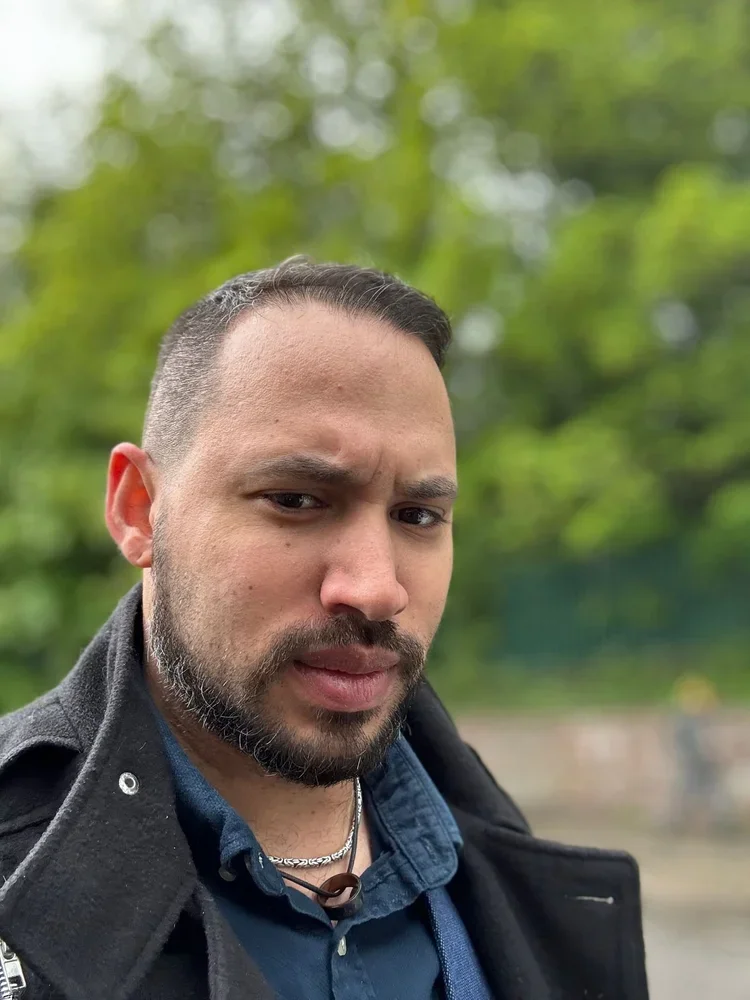 A man with short dark hair and a beard taking a selfie outdoors, with a blurry background of green trees.