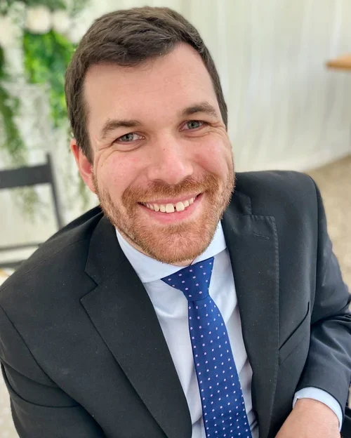 A smiling man with a beard in a black suit and blue polka dot tie, sitting indoors with a green plant in the background.