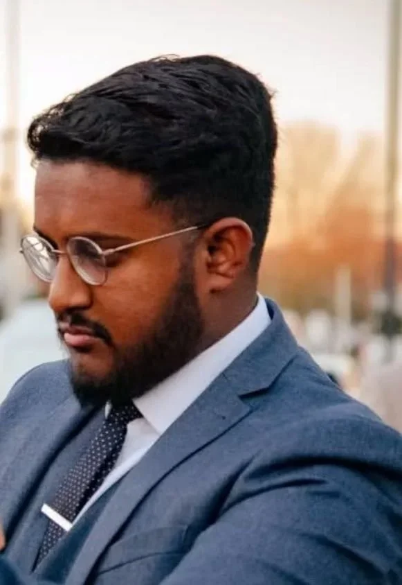 A man wearing a blue suit, white shirt, and black tie with white accents looking down, with glasses and a well-groomed beard, outdoors with blurry trees and buildings in the background.