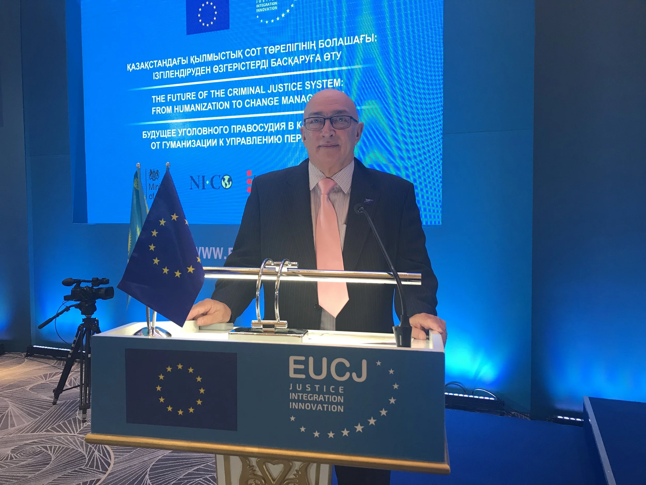Man in suit with pink tie speaking at a podium with European Union flag, with a blue-themed backdrop displaying event information in multiple languages, including English and Kazakh, about the future of the criminal justice system in Kazakhstan.