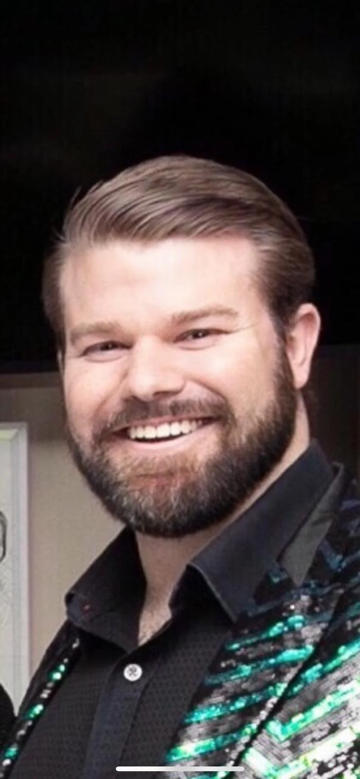 Smiling man with a beard and neatly combed hair, wearing a black shirt with shiny, colorful decoration on the shoulders.