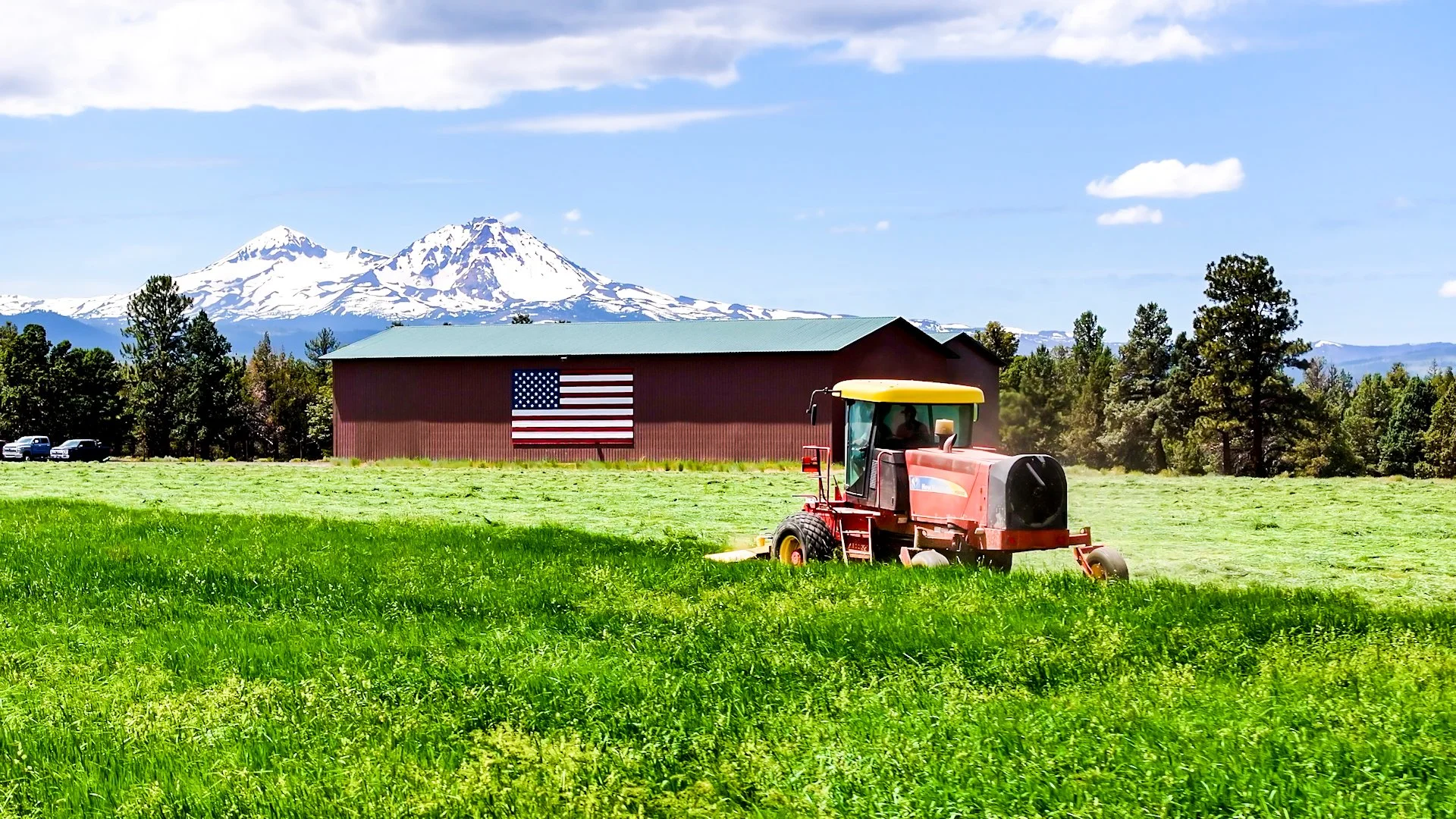 A red tractor working on a lush green field with a barn, displaying the American flag, and snow-capped mountains in the background under a partly cloudy sky.