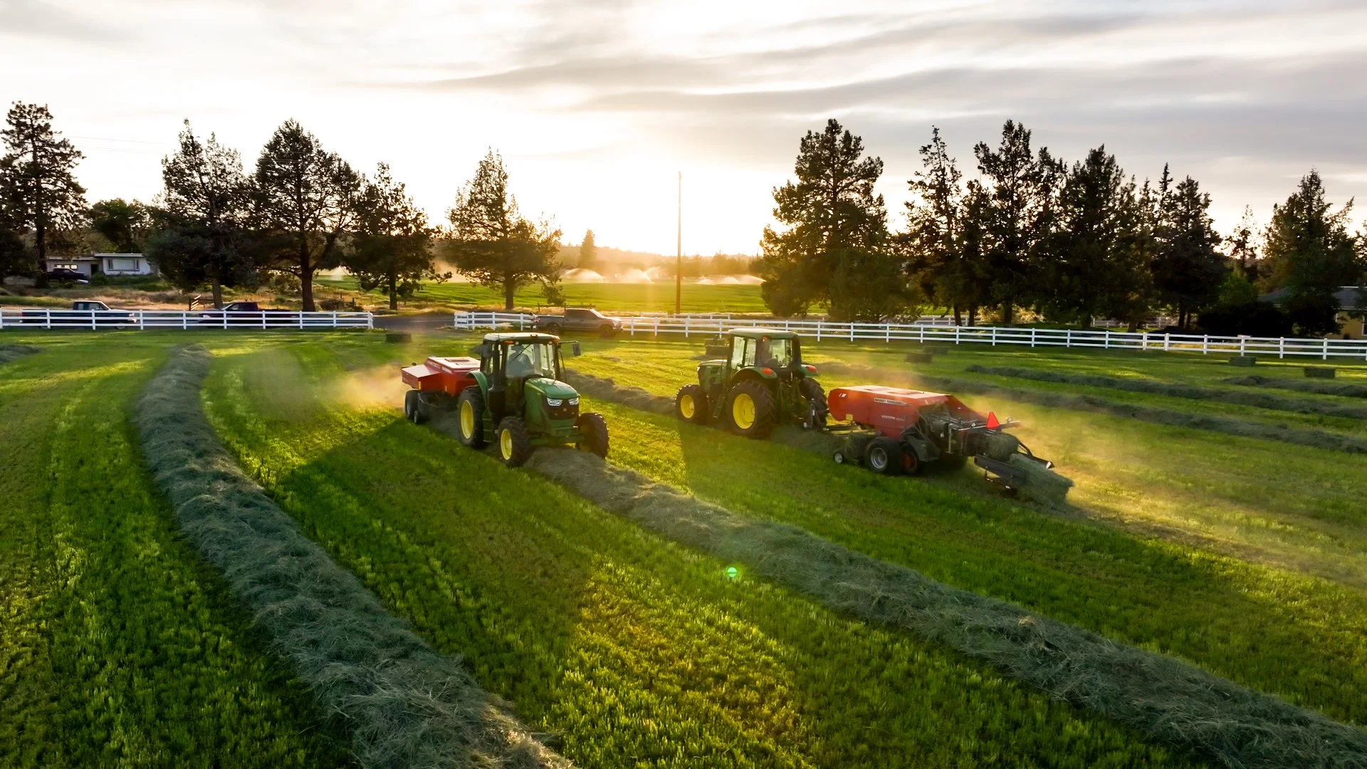 Two tractors mowing fields on a farm during sunset with trees, houses, and a white fence in the background.
