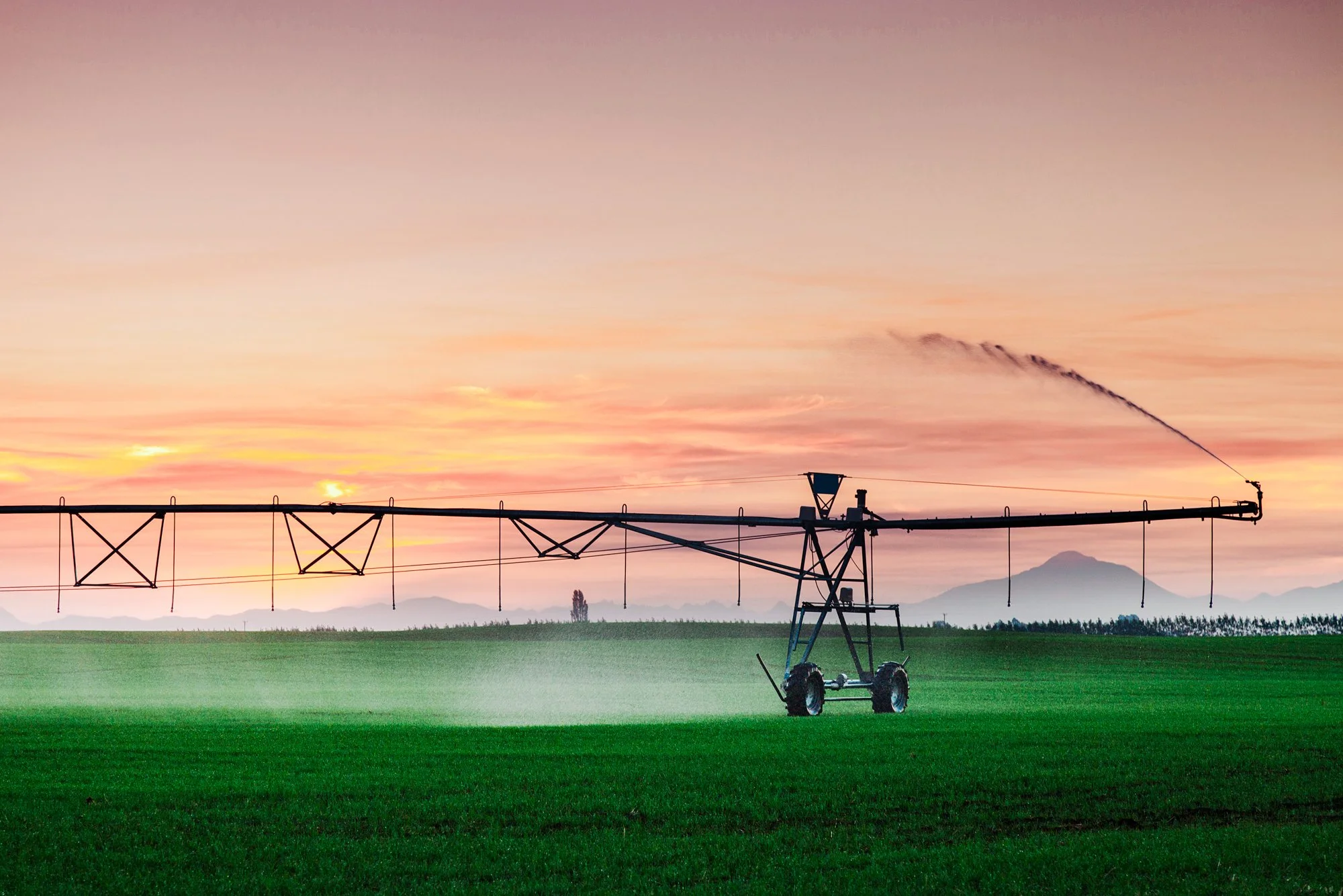 A large irrigation sprinkler spraying water over a green field at sunset with mountains in the background.