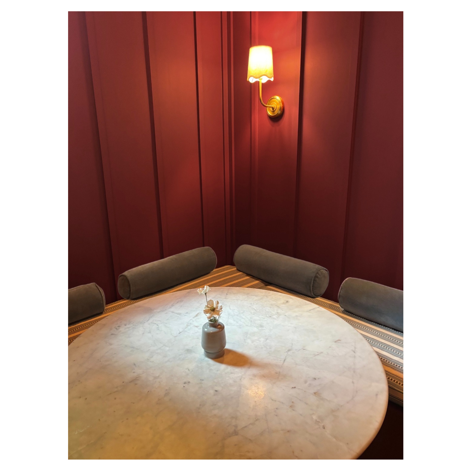 An intimate dining nook designed by Austin interior designer HOLLEYHOUSE with a round table set surrounded by a cushioned bench with striped fabric and a padded chair, against red paneled walls illuminated by wall sconces.