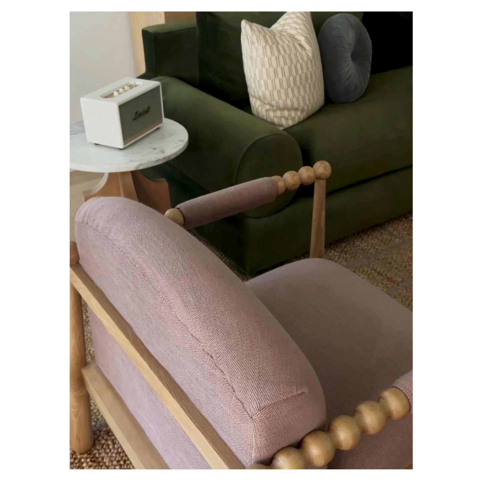 The image shows a living room with a pink upholstered armchair in the foreground, a dark green velvet sofa with coordinating pillows behind it, a small round marble side table with a white speaker on top, and a patterned rug on the floor.