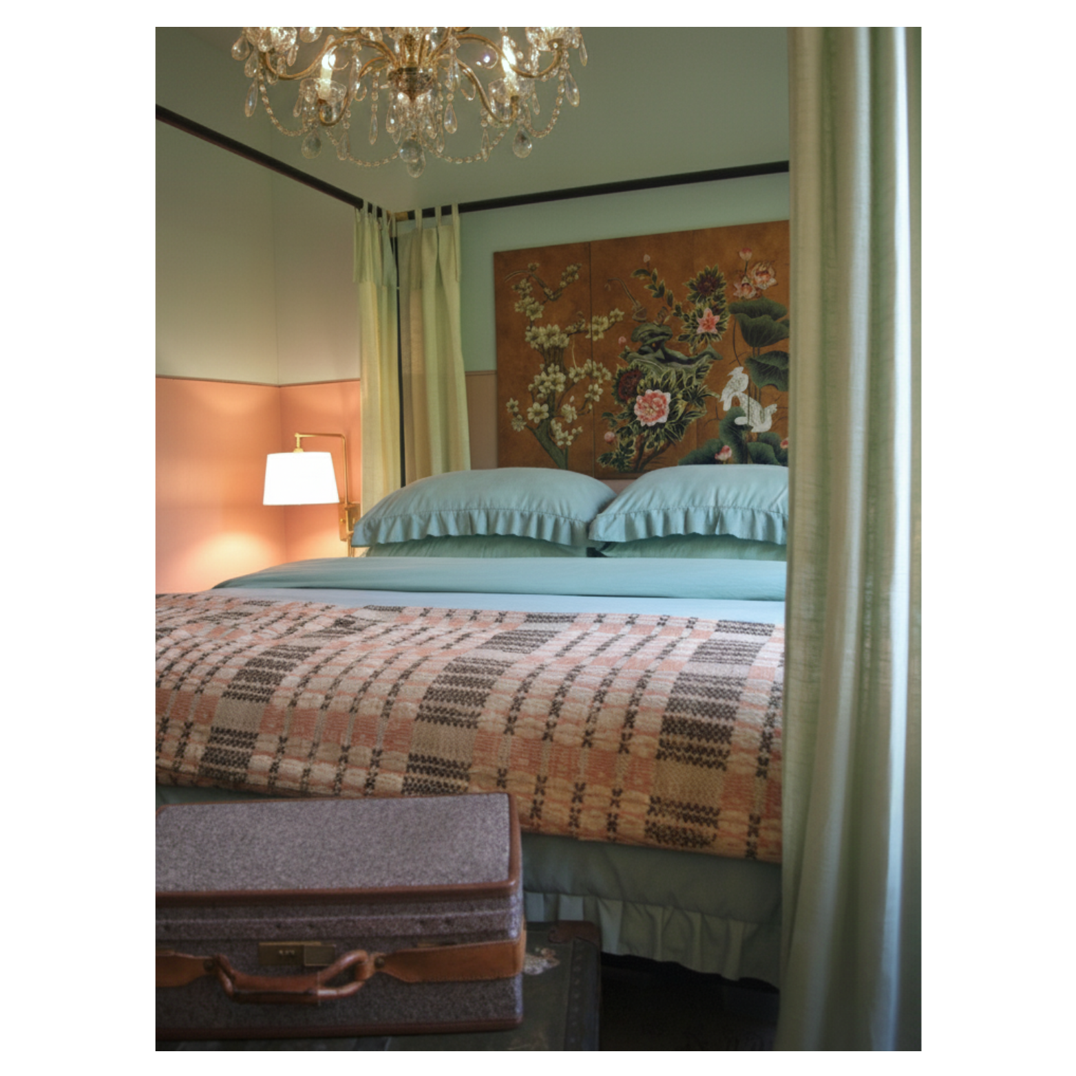 A hotel room featuring a bed with pastel blue sheets and ruffled pillows, a checkered blanket, and a floral-pinched headboard with Asian-style art above it. To the left, there is a wall-mounted lamp and a green curtain partially covering the window, with a chandelier hanging above.