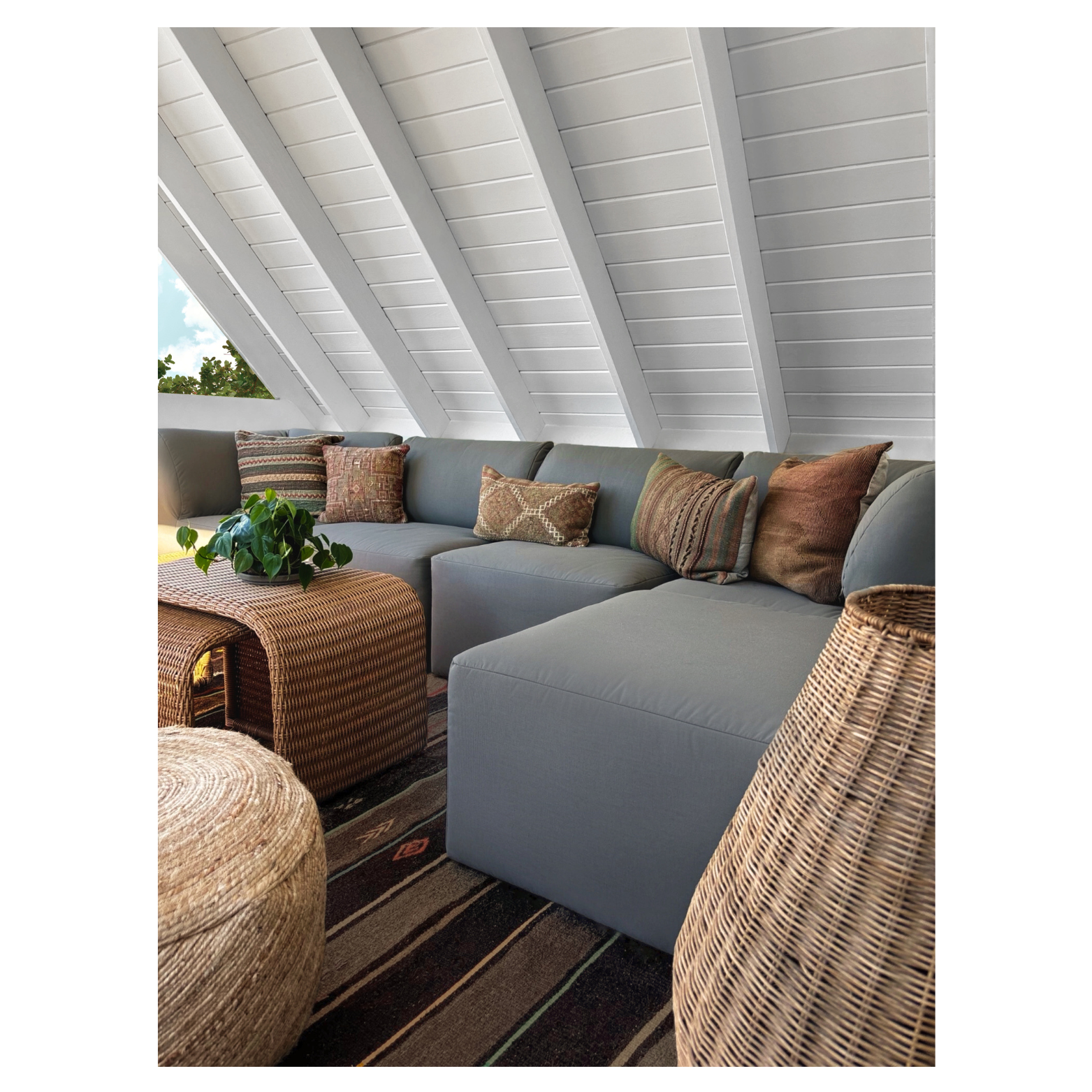 Cozy attic lounge area designed by Austin interior designer HOLLEYHOUSE with a gray sectional sofa with patterned cushions, a woven rattan coffee table with a green plant, and wicker ottomans on a striped rug under a white sloped ceiling.