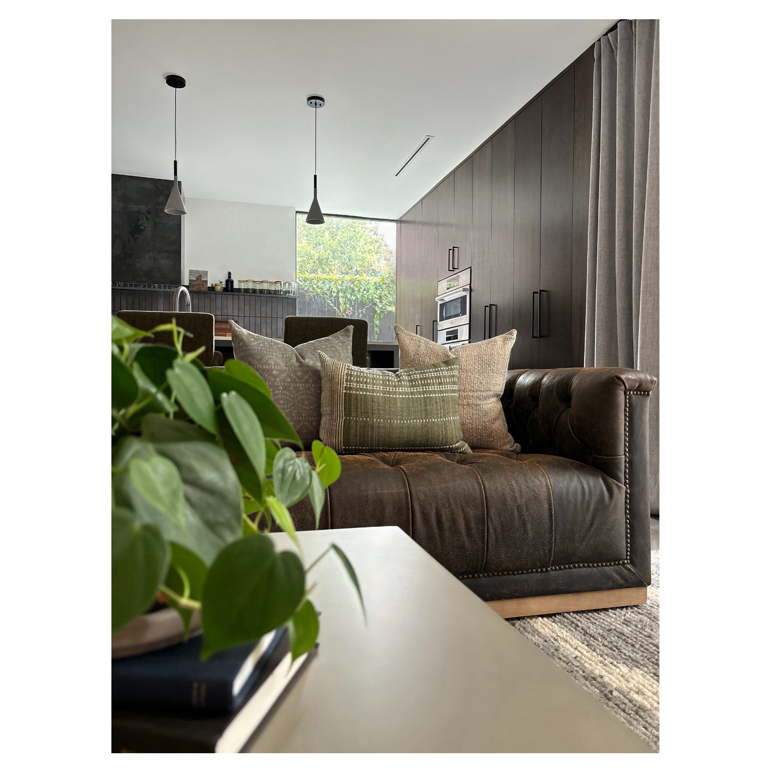 Living room designed by Austin interior designer HOLLEYHOUSE with brown leather tufted sofa, multiple throw pillows, a white coffee table with a plant and book, against a backdrop of a modern kitchen.