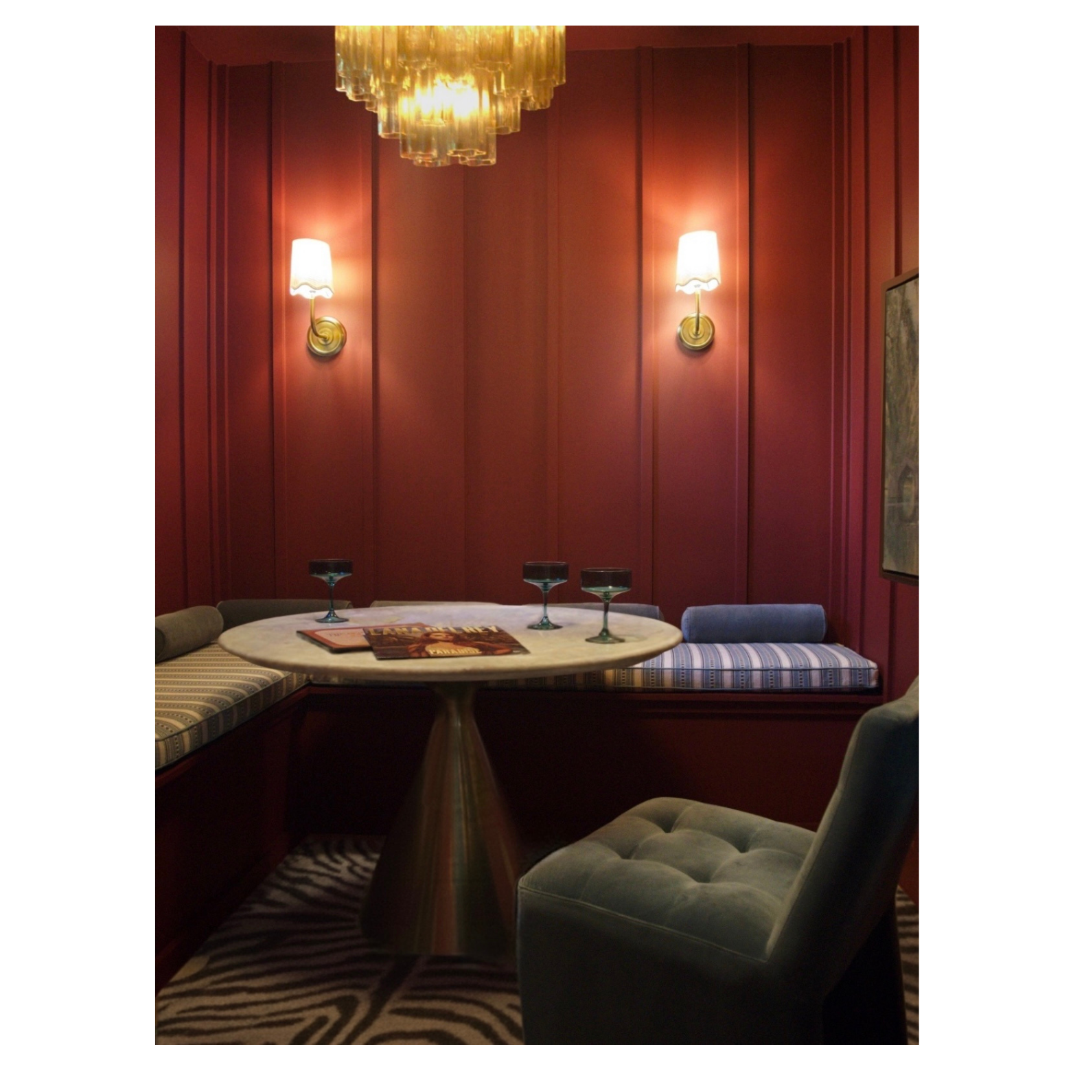 A cozy corner with a round marble table, a plush chair, built-in bench seating with striped cushions, wall sconces, a chandelier, and a zebra-print rug in a warmly lit room.