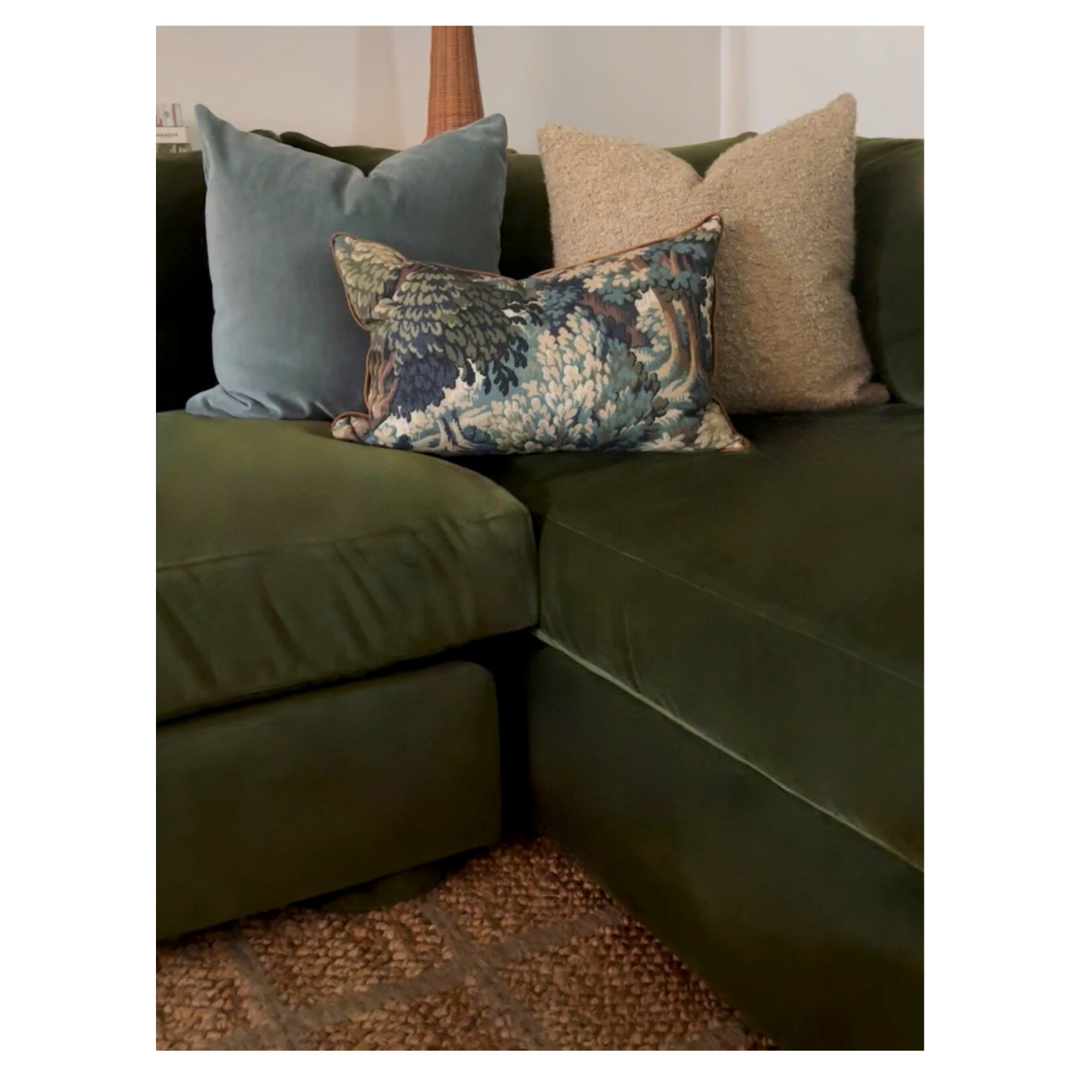 A green sectional sofa selected by HOLLEYHOUSE with four decorative pillows on it, including a floral embroidered pillow in the front, a beige textured pillow, a blue velvet pillow, and a wicker lamp in the background.